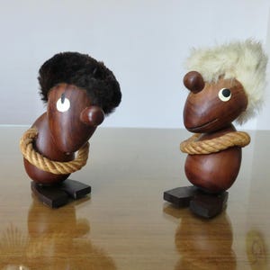 Può includere: Due statuette di legno con corpi marrone scuro, sezioni centrali a forma di corda e piedi marrone scuro. Uno ha una copertura per la testa simile a pelliccia marrone scuro, l'altro ha una copertura per la testa simile a pelliccia beige chiaro. Le statuette hanno occhi bianchi e nasi marroni.