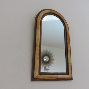 May include: A decorative arched mirror with a brown frame and a light brown, segmented border. The mirror reflects a white ceiling and a gold sunburst mirror.