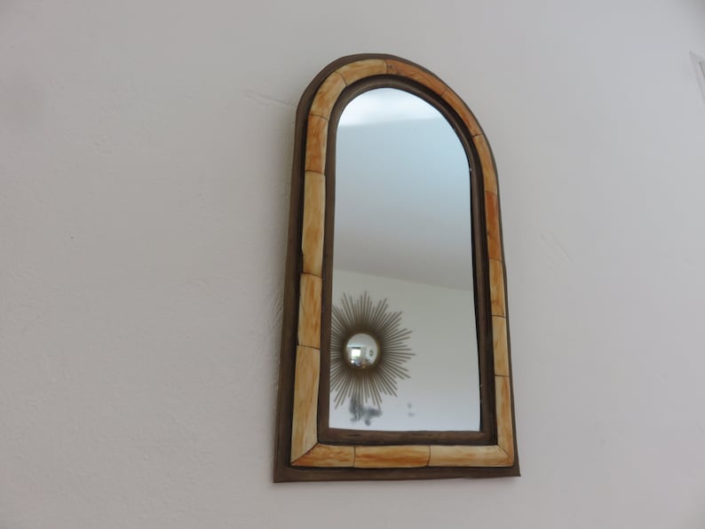 May include: A decorative arched mirror with a brown frame and a light brown, segmented border. The mirror reflects a white ceiling and a gold sunburst mirror.