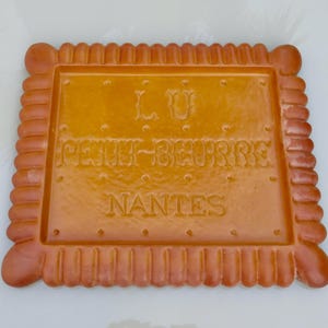 Könnte beinhalten: Ein quadratisches, goldbraunes Keramiktablett in Keksform mit gewelltem Rand und abgerundeten Ecken. In der Mitte befindet sich ein geprägter Schriftzug mit LU, PETIT-BEURRE und NANTES, umgeben von einem Raster aus kleinen, kreisförmigen Vertiefungen.