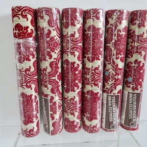 Könnte beinhalten: Sechs Rollen Tapete mit Muster. Die Tapete hat einen cremefarbenen Hintergrund mit einem sich wiederholenden roten Blumendesign. Die Rollen sind in Klarsichtfolie verpackt und mit "VELOURS FLOCK" beschriftet.