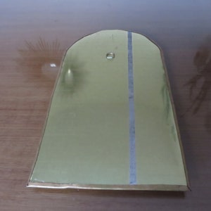 May include: A gold-colored metal plaque with a rounded top and a vertical line down the center. The plaque has a small hole near the top.