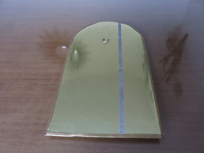 May include: A gold-colored metal plaque with a rounded top and a vertical line down the center. The plaque has a small hole near the top.