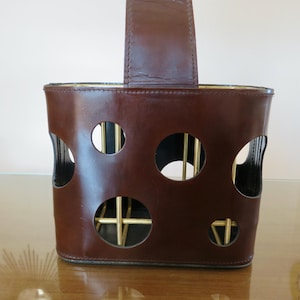 May include: A brown leather wine bottle holder with a handle. The holder features circular cutouts and gold-coloured metal supports inside. The handle is made of the same brown leather. The holder is on a reflective surface.