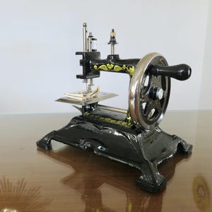 Könnte beinhalten: Eine schwarze Vintage-Nähmaschine mit goldenen floralen Akzenten. Die Maschine hat ein großes silbernes Schwungrad und eine kleine Stichplatte. Der Sockel ist verziert, und die Maschine scheint gut erhalten zu sein.