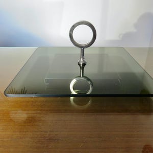 Puede incluir: Una bandeja cuadrada de vidrio con un asa plateada. La bandeja es de color gris ahumado y se encuentra sobre una superficie de madera. El asa es un anillo circular unido a un pedestal.