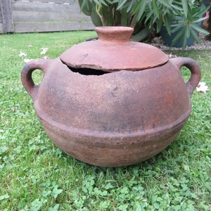 Puede incluir: Una olla grande de barro marrón con tapa. La olla tiene dos asas y una tapa astillada. La olla está sentada sobre un lecho de hierba verde.