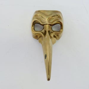 May include: A gold-coloured metal mask with a long, pointed nose and two eye holes. The mask has a textured surface and appears to be made of brass.