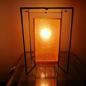 May include: A rectangular table lamp with a warm orange glow. The lamp has a black metal frame and a rectangular shade that diffuses the light. The lamp is on a glass surface.