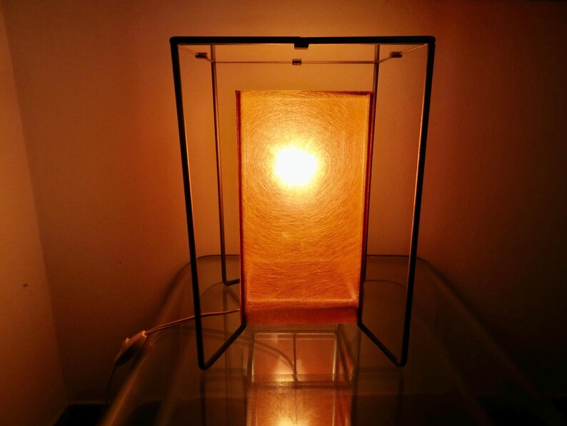 May include: A rectangular table lamp with a warm orange glow. The lamp has a black metal frame and a rectangular shade that diffuses the light. The lamp is on a glass surface.