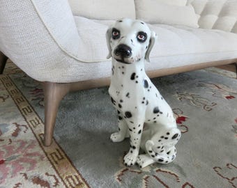 May include: A ceramic Dalmatian dog figurine, white with black spots, sits on a patterned rug. The dog has brown eyes and a black nose. The figurine is near a light-colored sofa with wooden legs.
