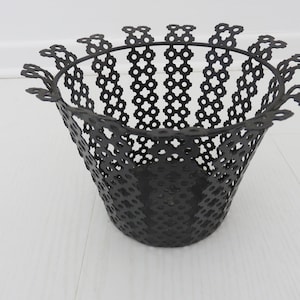 Könnte beinhalten: Schwarzer Metalltopf mit einem dekorativen, gewellten Rand und einem geometrischen Muster. Der Topf ist aus Drahtgeflecht und hat eine konische Form.