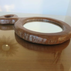 Puede incluir: Un espejo de mano de madera con un espejo redondo y un mango único. El espejo está rodeado por un marco de madera grueso y tallado.