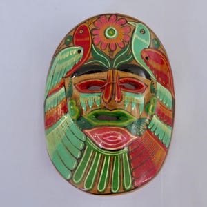 Peut inclure: Masque ovale en bois peint à la main avec un visage. Le masque présente un visage central avec des détails rouges, verts et noirs, flanqué d'oiseaux rouges et verts. Une fleur est en haut. C'est un objet décoratif.