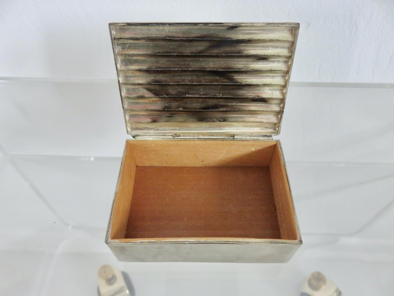 Pu&ograve; includere: Una scatola di metallo argentato con interno in legno. La scatola ha un design a coste sul coperchio ed &egrave; aperta per rivelare l'interno vuoto.