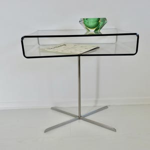 May include: A modern side table with a clear glass top and a lower shelf. The table has a black trim and a single, silver-colored leg with a star-shaped base. A green glass bowl and a piece of paper rest on the table.