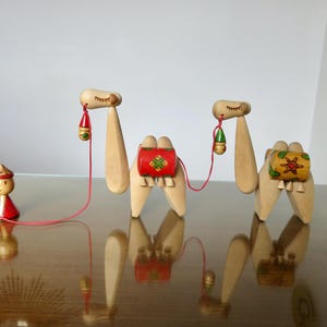 Op de afbeelding: Drie houten kamelenfiguren met kleurrijke gepatroneerde zadels en een klein houten figuurtje van een persoon met een rode hoed en een gewaad. De kamelen zijn verbonden door een rood touw.