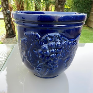 May include: A cobalt blue ceramic planter with a glossy finish. The planter features a raised relief design of grape clusters and leaves. The pot has a wide, rounded shape and is suitable for outdoor or indoor use.