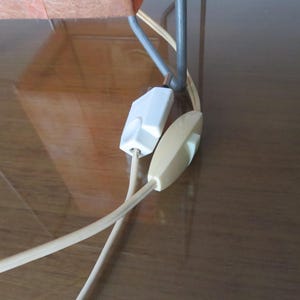 May include: Close-up of a lamp cord featuring a white inline switch and a cream-coloured on/off switch. The beige cord connects to a white plug. The lamp's metal frame is visible in the background.