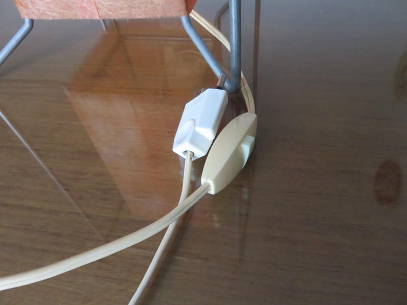 May include: Close-up of a lamp cord featuring a white inline switch and a cream-coloured on/off switch. The beige cord connects to a white plug. The lamp's metal frame is visible in the background.