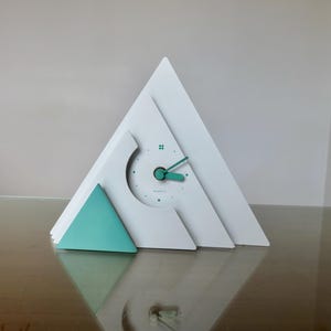 Puede incluir: Un moderno reloj de cuarzo blanco con un diseño geométrico. El reloj presenta una forma triangular con un acento turquesa y manecillas turquesas. La esfera es blanca con puntos negros y la palabra "QUARTZ".