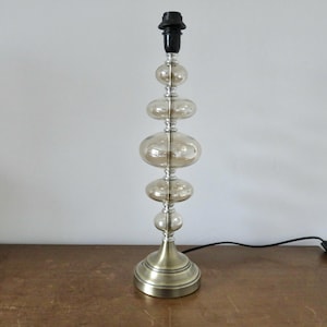 Könnte beinhalten: Dekorative Tischlampe mit einem Design aus übereinander angeordneten, kugelförmigen Elementen. Die Lampe hat einen messingfarbenen Sockel und Akzente, mit klaren Glaskugeln in einem hellen Bernsteinton. Sie hat eine schwarze Lampenfassung und ein schwarzes Netzkabel.