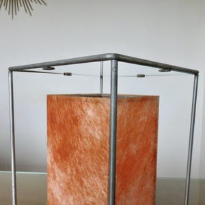 May include: A contemporary lamp featuring an orange, textured rectangular shade within a grey metal frame. The lamp's design is minimalist, with a simple, geometric structure. The shade appears to be made of a fibrous material.