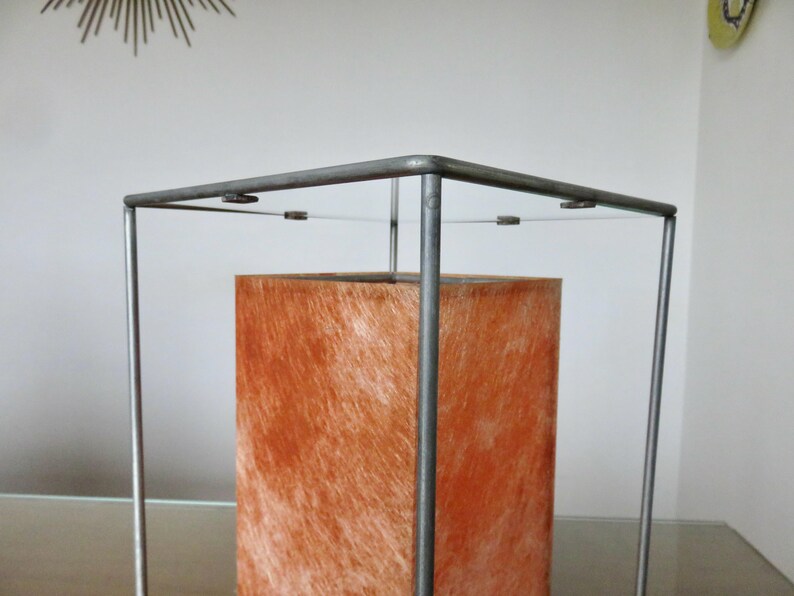 May include: A contemporary lamp featuring an orange, textured rectangular shade within a grey metal frame. The lamp's design is minimalist, with a simple, geometric structure. The shade appears to be made of a fibrous material.