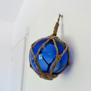 Può includere: Una boa di vetro blu, sospesa da una rete di corda naturale e un anello di corda ritorta. La boa è di un colore blu vibrante e pende contro un muro bianco, suggerendo un arredamento nautico.