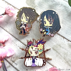 May include: Three gold-coloured enamel pins featuring anime-style characters. The first pin features a male character with black hair and a white and black outfit. The second pin features a female character with long black hair and a blue and white outfit. The third pin features a female character with blonde hair and a purple and gold outfit.