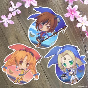 Peut inclure: Trois autocollants de personnages de style anime. Chaque autocollant présente un personnage de style chibi avec des tenues et des accessoires uniques. Les autocollants sont placés sur un fond circulaire avec un ciel et un design de lune. Les autocollants sont sur une surface en bois.