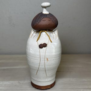 Puede incluir: Figura de cerámica hecha a mano de una persona con cabeza marrón y cuerpo blanco. La cabeza tiene los ojos cerrados y una pequeña sonrisa. El cuerpo es blanco con detalles marrones y una cinta amarilla. La figura mide unos 20 cm de altura.