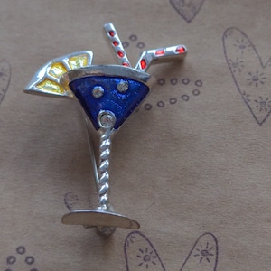 May include: A silver-toned cocktail glass brooch featuring a deep blue enamel bowl with three embedded clear stones. The glass is garnished with a yellow enamel citrus slice and contains a red enamel striped straw. The stem has a twisted design.