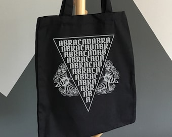 May include: Black tote bag with the word "ABRACADABRA" in a white triangle design. The bag has black handles and is hanging on a wooden coat rack. The background is a gray and white wall.