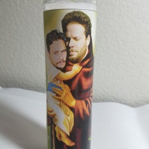 May include: A glass candle with a printed image of two men. The man on the left is holding a child. The man on the right is wearing a brown robe. The candle is white and has a green wick.