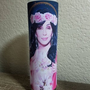 May include: A glass candle with a white candle inside. The candle has a label with a picture of a woman with a flower crown on her head. The woman is wearing a pink dress and has her hands clasped in front of her chest.