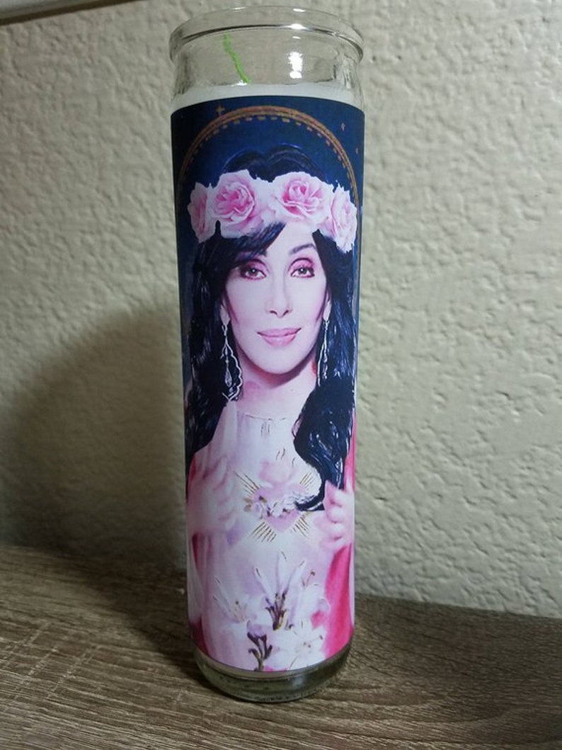 May include: A glass candle with a white candle inside. The candle has a label with a picture of a woman with a flower crown on her head. The woman is wearing a pink dress and has her hands clasped in front of her chest.