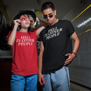 May include: Two people wearing t-shirts with the text "HELL IS OTHER PEOPLE" in white. One shirt is red, the other is black. The person on the left is wearing a black hat and red sunglasses. The person on the right is wearing sunglasses.