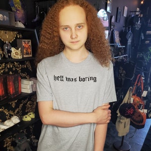 Puede incluir: Una camiseta gris claro con la frase "hell was boring" en negro con fuente gótica. La camiseta es de cuello redondo. La persona de la imagen tiene el pelo rizado rojo.