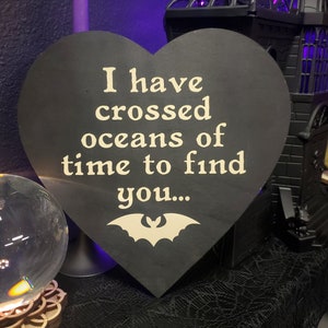 Könnte beinhalten: Schwarzes herzförmiges Holzschild mit dem cremefarbenen Text "I have crossed oceans of time to find you...". Eine cremefarbene Fledermausgrafik befindet sich unten. Das Schild ist Teil eines Halloween- oder Gothic-Themas.
