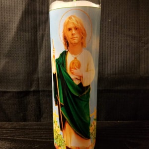 May include: A glass candle with a white wax base and a printed image of a man with long blonde hair. The image is of a man in a green robe with a gold necklace and a gold heart pendant. The candle is on a dark wooden surface.