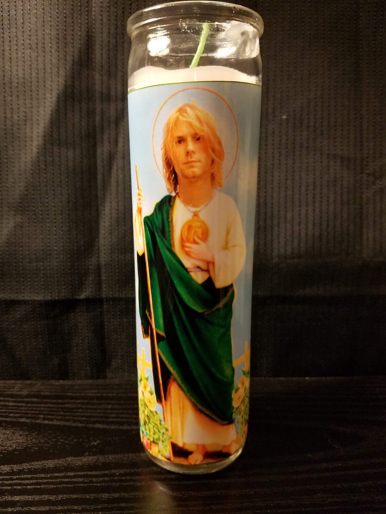 May include: A glass candle with a white wax base and a printed image of a man with long blonde hair. The image is of a man in a green robe with a gold necklace and a gold heart pendant. The candle is on a dark wooden surface.