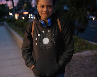 Puede incluir: Sudadera negra con un gráfico blanco de un sol y las fases lunares. La sudadera tiene un bolsillo delantero y una capucha con cordón. La persona de la imagen lleva unos auriculares azules alrededor del cuello.