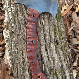 May include: A handmade axe with a dark gray metal head and a red wood handle. The handle has a textured design and brass accents.
