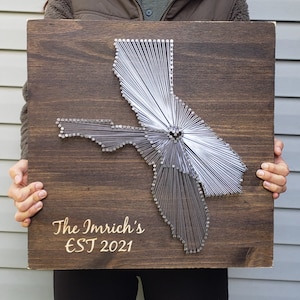 May include: A wooden wall art piece featuring a string art design of the California and Florida state outlines. The dark brown stained wood board has the text "The Imrich's EST 2021" in a gold color.