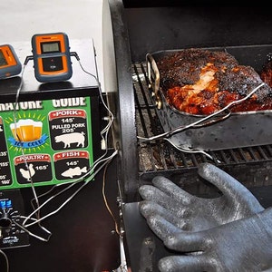 May include: Two large cuts of meat are being smoked in a black smoker. The meat is in a metal pan and has a dark brown crust. A digital thermometer is attached to the meat. A temperature guide is on the side of the smoker with a chart for different types of meat and their recommended cooking temperatures.