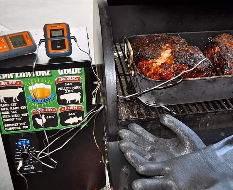 May include: Two large cuts of meat are being smoked in a black smoker. The meat is in a metal pan and has a dark brown crust. A digital thermometer is attached to the meat. A temperature guide is on the side of the smoker with a chart for different types of meat and their recommended cooking temperatures.