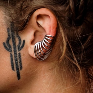 Puede incluir: Un brazalete de plata con un diseño en capas se usa en la oreja. Un tatuaje negro es visible en el costado de la cara. La persona lleva una camiseta negra. El cabello es castaño y ondulado.