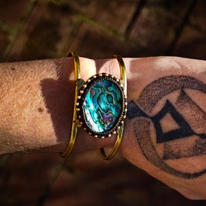 Könnte beinhalten: Ein doppeltes Armreif-Armband in Goldton mit einem großen ovalen Einlegeteil aus Abalone-Muschel. Die Muschel hat ein blaues, grünes und violettes irisierendes Muster.