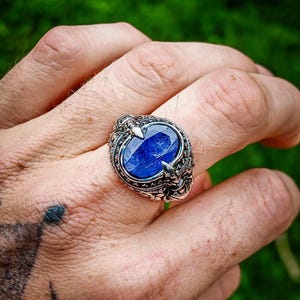 Peut inclure: Une bague en argent ornée d'une pierre précieuse ovale d'un bleu profond, montée sur une bande métallique détaillée et travaillée. Le sertissage présente des motifs en relief complexes entourant la pierre centrale facettée.